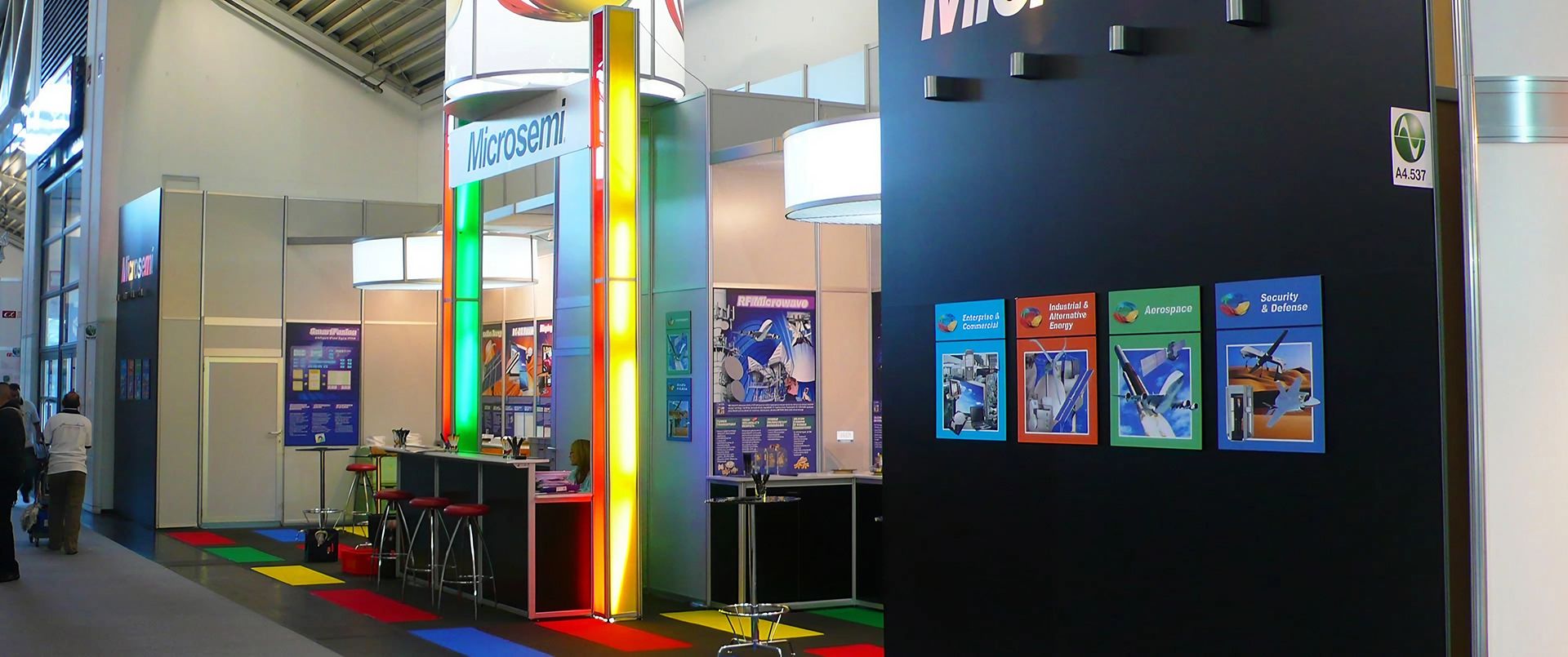Messestand mit bunten Leuchtstreifen, Barhockern und Informationsplakaten in einer Halle